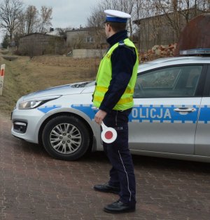 Policjant umundurowany w żółtej kamizelce i białej czapce stoi przy radiowozie zaparkowanym przy drodze