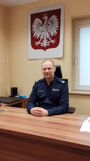 umundurowany policjant siedzi za biurkiem, za nim na ścianie wisi godło Polski