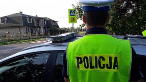 Policjant umundurowany w białej czapce na głowie stoi odwrócony tyłem przed radiowozem i patrzy na drogę