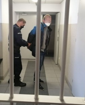 umundurowany policjant zakłada zatrzymanemu mężczyźnie kajdanki na ręce z tyłu, stoją obaj za zakratowanymi drzwiami