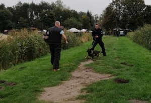 umundurowany policjant trzyma przed sobą na smyczy psa, który szczeka na mężczyznę idącego w jego kierunku