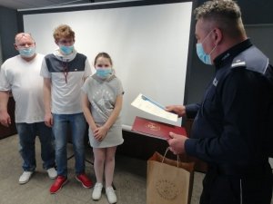 umundurowany policjant wręcza dziewczynce dyplom i prezent, obok dziewczynki stoi dwóch mężczyzn, na twarzach wszyscy mają maseczki