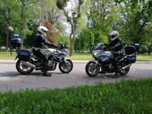 Dwaj umundurowani policjanci w białych kaskach na głowie jadą drogą siedząc na policyjnych motocyklach