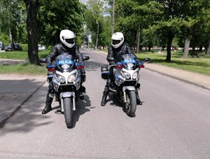 Dwaj umundurowani policjanci w białych kaskach na głowie jadą drogą siedząc na policyjnych motocyklach