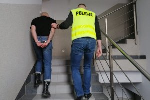 klatka schodowa w komendzie, policjant w żółtej kamizelce z napisem policja prowadzi zatrzymanego mężczyznę