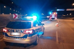 radiowóz policyjny z migającymi niebieskimi światłami stoi na miejscu wypadku drogowego, w tle widoczny wóz strażacki