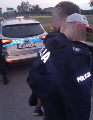 Mężczyzna trzyma na rękach chłopca w czapeczce na głowie, chłopiec okryty jest bluzą z napisem policja, za nimi stoi radiowóz policyjny