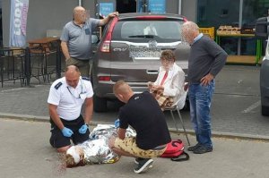 umundurowany policjant udziela pomocy leżącej kobiecie, policjant jest schylony przy kobiecie wraz z innym mężczyzną który mu pomaga. Obok siedzi na krzesełku inna kobieta i stoi dwóch mężczyzn