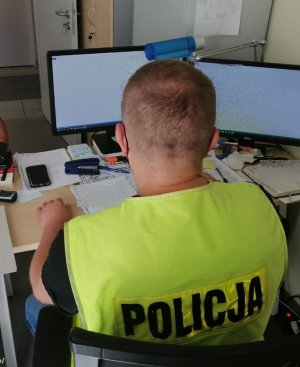nieumundurowany policjant w żółtej kamizelce z napisem policja siedzi odwrócony tyłem, patrzy w monitor komputera