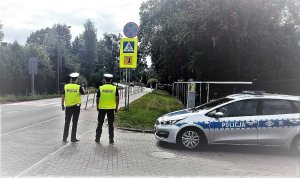 umundurowani policjanci w białych czapkach na głowie i żółtych kamizelkach z napisem policja stoją przy drodze odwróceni tyłem, obok nich stoi zaparkowany radiowóz
