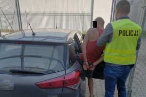 policjant w żółtej kamizelce z napisem policja na plecach prowadzi zatrzymanego mężczyznę, który ma spięte ręce kajdankami