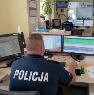 dwóch policjantów w granatowych mundurach siedzi za biurkami, są odwróceni tyłem. Siedzą przed stojącymi na blatach komputerami, przed nimi jest duża szyba w ścianie przez którą widoczny jest duży niebieski napis Policja