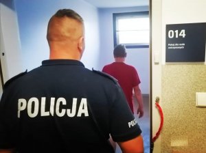 policjant w granatowym mundurze odwrócony tyłem na plecach ma napis policja, stoi w drzwiach pomieszczenia gdzie stoi odwrócony tyłem zatrzymany mężczyzna w czerwonej koszulce