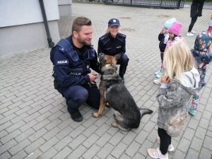 umundurowany policjant kuca przy psie, obok niego umundurowana policjantka i wokół nich stoją dzieci