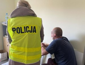 policjantka w żółtej kamizelce z napisem policja stoi przy zatrzymanym mężczyźnie, który siedzi przy stoliku z kajdankami na rękach