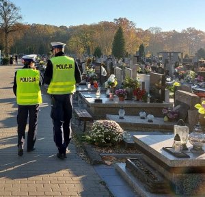 umundurowany policjant i policjantka w żółtych kamizelkach z napisem policja idą alejką cmentarza, wokół są groby