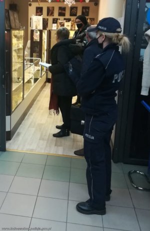 umundurowana policjantka w maseczce na twarzy stoi bokiem przy wejściu do sklepu, w którym przy ladzie stoją 3 osoby z założonymi maseczkami na twarzy