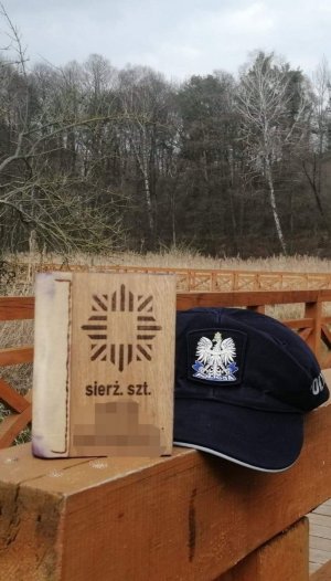Pudełko drewniane z symbolem policyjnym i napisem sierż. szt. obok położona policyjna granatowa czapka, w tle widać duże drzewa