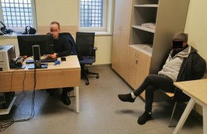 umundurowany policjant siedzi za biurkiem przed monitorem komputera, po prawej stronie fotografii siedzi na krześle zatrzymany mężczyzna