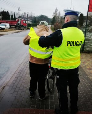Dwaj umundurowani policjanci w żółtych kamizelkach i białych czapkach na głowie dawali rowerzystom kamizelki odblaskowe i pomagali im je założyć