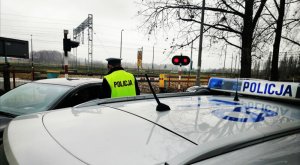 policjant w białej czapce na głowie i w żółtej kamizelce z napisem policja stoi odwrócony tyłem, stoi przy samochodzie i rozmawia z kierowcą, za nimi jest przejazd kolejowy