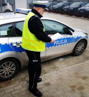 Umundurowany policjant w białej czapce na głowie i w żółtej kamizelce stoi przy zaparkowanym radiowozie i patrzy w dokumenty trzymane w dłoniach