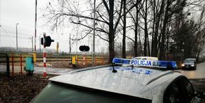 radiowóz policyjny stoi przy przejeździe kolejowym , gdzie są opuszczone zapory i miga sygnalizator na czerwono