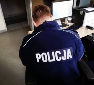 policjant w granatowym mundurze z białym napisem na plecach policja siedzi odwrócony tyłem przy biurku