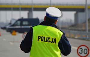 policjant w żółtej kamizelce z napisem policja na plecach stoi odwrócony tyłem , na głowie ma białą czapkę, w tle zablokowana droga