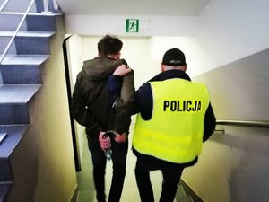 policjant w żółtej kamizelce z napisem policja schodzi po schodach z zatrzymanym mężczyzną i trzyma go pod ramię