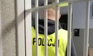 policjant w żółtej kamizelce z napisem policja stoi odwrócony tyłem za okratowanymi drzwiami