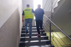 Policjant w żółtej kamizelce prowadzi po schodach zatrzymanego mężczyznę