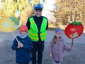 Dwóch policjantów w żółtych kamizelkach stoi wraz z dziećmi i nauczycielami z transparentami przedstawiającymi jabłko i cytrynę