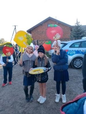 trzy dziewczynki stoją przed budynkiem, jedna z nich w środku trzyma tacę z cytryną a po bokach jej dziewczynki trzymają transparenty symbolizujące jabłko i cytrynę