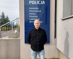 Policjant stoi przed budynkiem komendy, gdzie umieszczony jest napis na niebieskim szyldzie z białymi napisami Policja Komenda Powiatowa Policji powiatu łódzkiego wschodniego