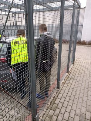 za okratowaną ścianą stoi przy samochodzie policjant w żółtej kamizelce i zatrzymany mężczyzna