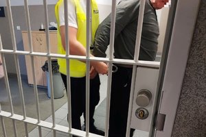 policjant w żółtej kamizelce zakłada kajdanki na ręce zatrzymanemu mężczyźnie, obaj stoją za okratowanymi drzwiami