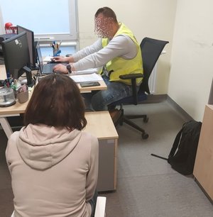 Policjant w żółtej kamizelce siedzi przy biurku na którym stoi komputer, obok niego siedzi zatrzymana kobieta
