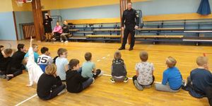 policjant stoi na środku sali gimnastycznej i mówi do dzieci siedzących przed nim na podłodze