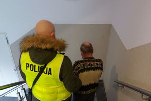 Dwóch policjantów, jeden z nich w żółtej kamizelce z napisem policja prowadzi zatrzymanego mężczyznę.