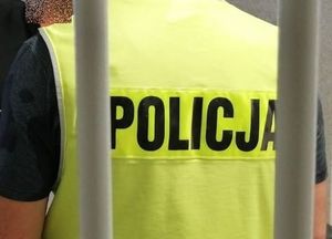 Policjant w żółtej kamizelce z napisem policja na plecach stoi za okratowanymi drzwiami.