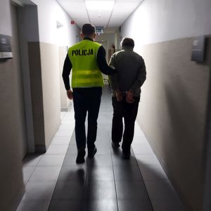 Policjant w żółtej kamizelce z napisem policja prowadzi przez korytarz zatrzymanego mężczyznę.