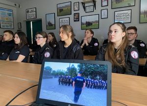 Uczniowie klas mundurowych siedzą w ławkach i patrzą przed siebie na rzutnik, na którym wyświetlany jest film ze stojącymi w dwuszeregu policjantami