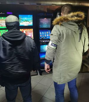 Dwóch mężczyzn stoi odwróconych tyłem, stoją przy automatach do gier, jeden z nich w zielonej kurtce ma założoną opaskę z napisem policja na lewym przedramieniu.