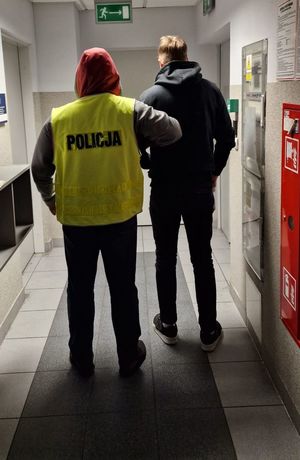 Mężczyzna w żółtej kamizelce z napisem policja i czerwonym kapturze na głowie prowadzi zatrzymanego mężczyznę.