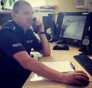 Policjant w mundurze, siedzi przy biurku na którym stoją ustawione monitory komputera, policjant trzyma przy uchu słuchawkę do telefonu.