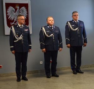 Trzech policjantów stoi w rzędzie obok siebie, przy ścianie, na ścianie wisi godło Polski.