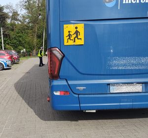 Policjant w żółtej kamizelce z napisem policja i białej czapce stoi przy dużym autobusie szkolnym w kolorze niebieskim.