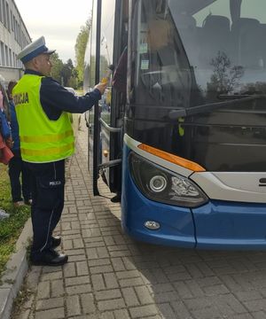 Policjant w białej czapce i żółtej kamizelce stoi przed otwartymi drzwiami autobusu i trzyma w dłoni żółte urządzenie do badania trzeźwości, które wyciąga w kierunku kierowcy autobusu.