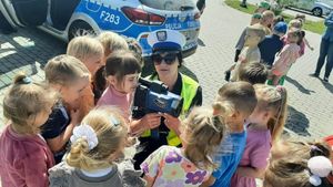 Policjantka kuca przy grupie małych dzieci i pokazuje im urządzenie do mierzenia prędkości.